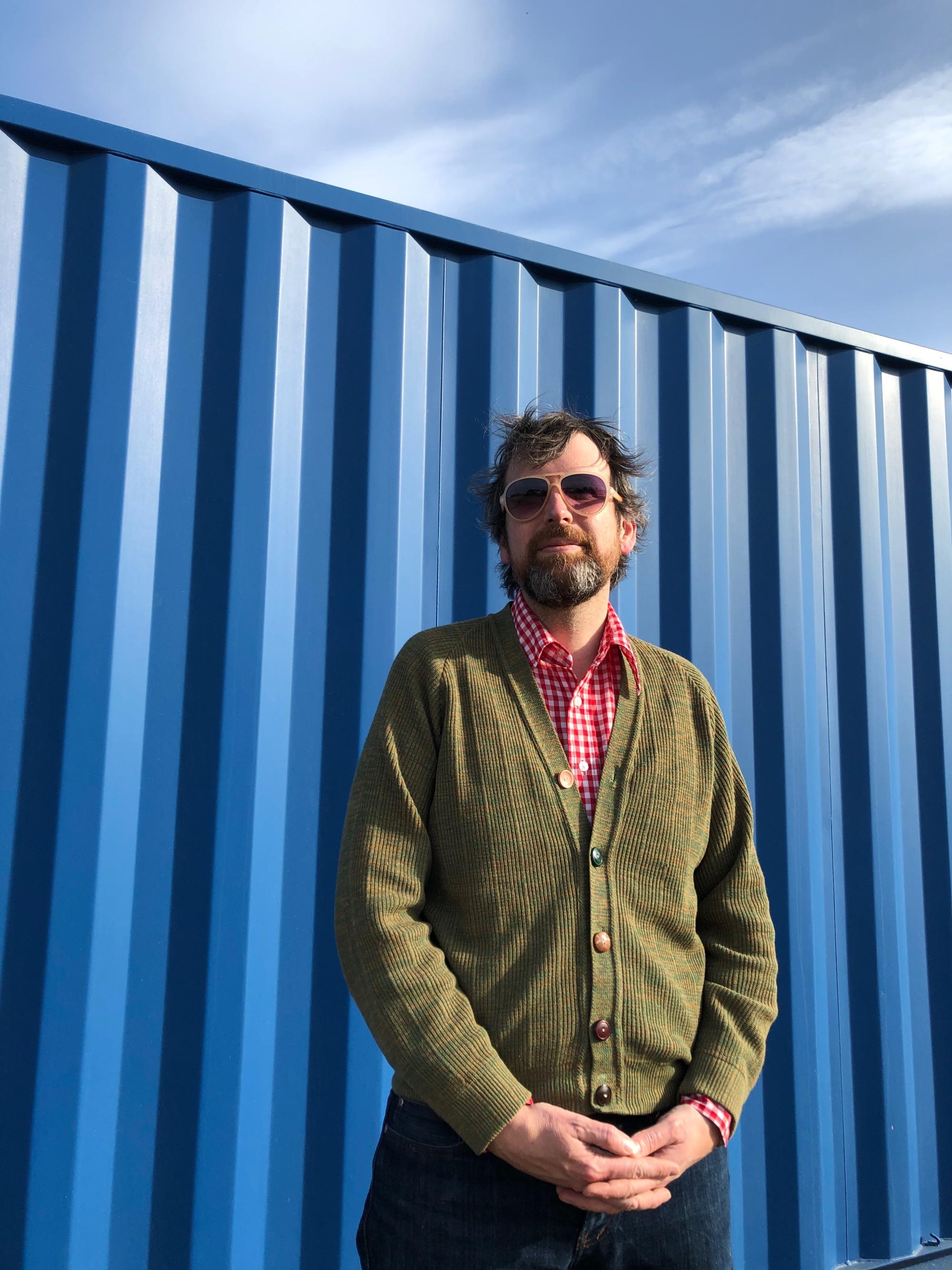 Jimmie stands in front of a blue shipping container, with light brown and gray hair and beard. He is wearing sunglasses, a red button-up, and olive green button up sweater