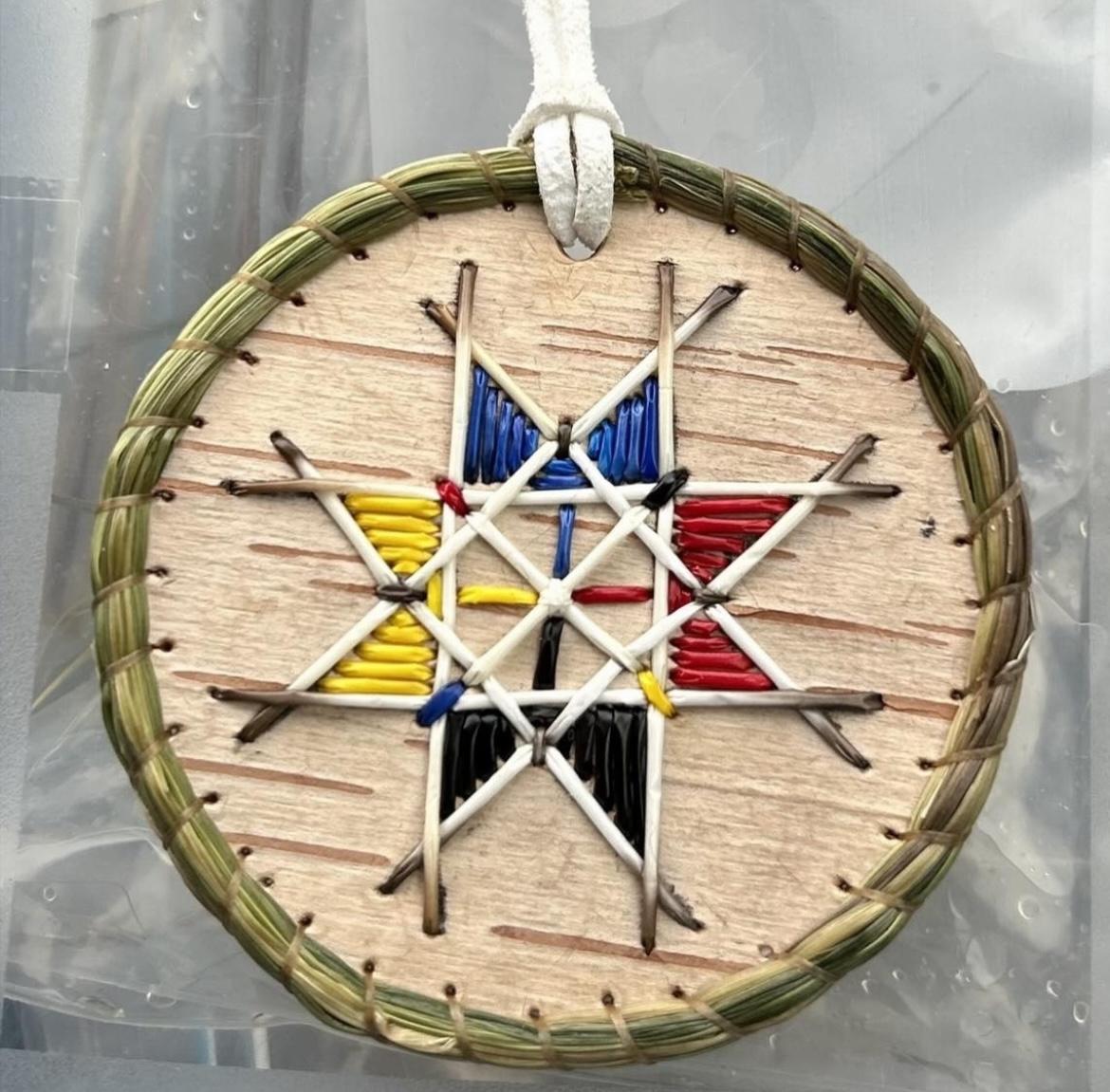 Photograph of a porcupine quillwork medallion made by Crystal Gloade on a round piece of birchbark and edged with sweetgrass. The quills make up a Mi'kmaq 8-point star design and are yellow, blue, red, black, and white.