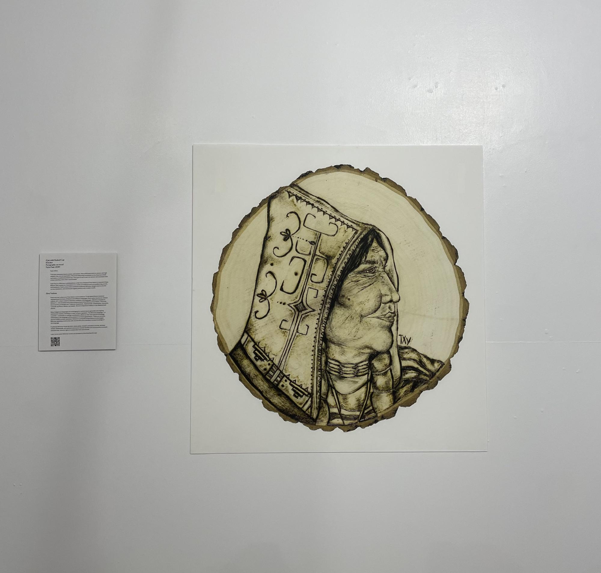 [ID: "circular disk of wood with woodburned illustration on the face of it depicting the side profile of an older l'nu woman wearing a peaked cap. She looks off to the right and is adorned with double curve motifs and bone jewellery."]