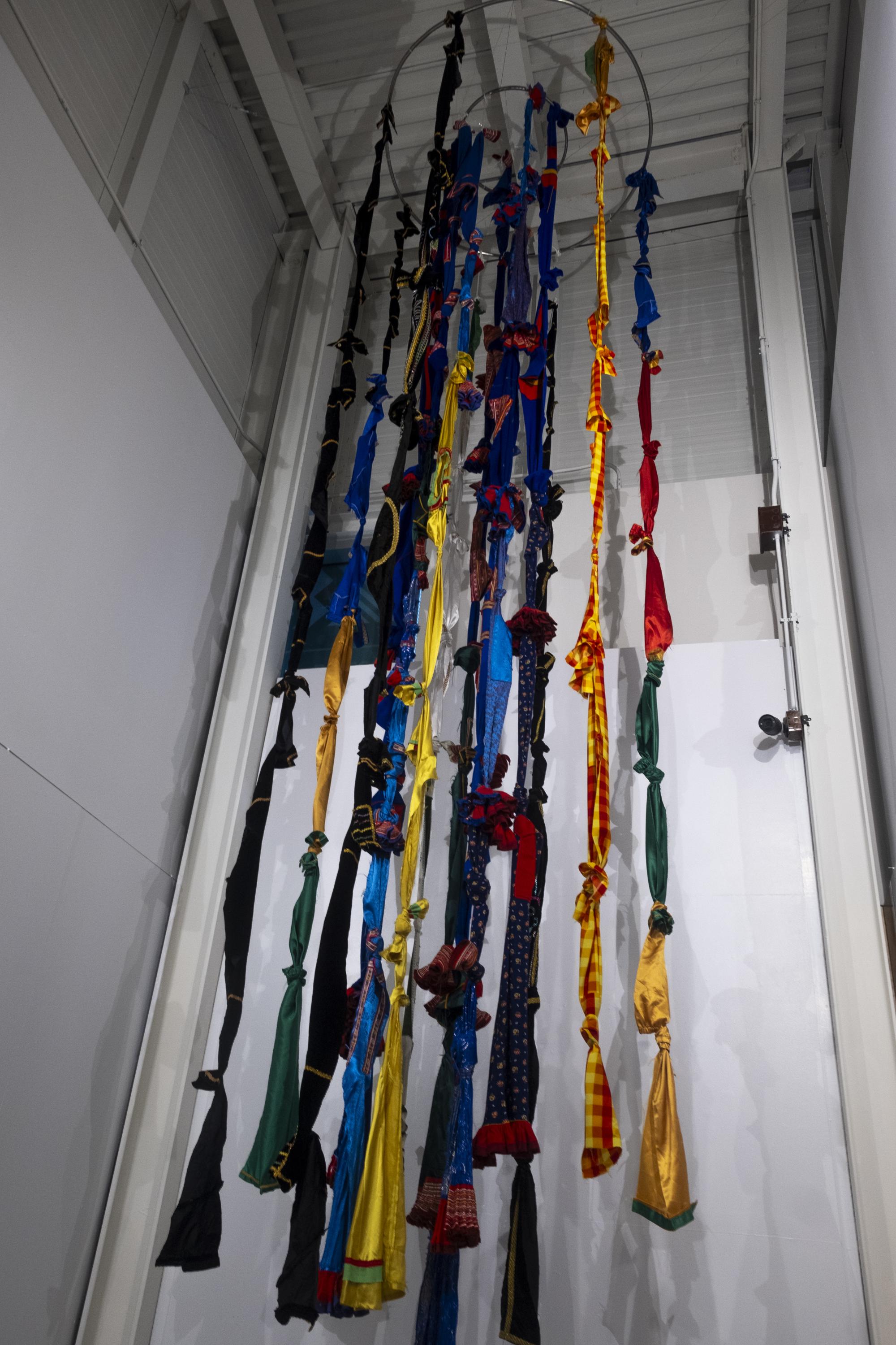 [ID: "A full length shot of the textile installation hanging from the ceiling from 2 metal hoops. Hanging from the hoops are long strips of colourful fabric tied with knots to form a quipo.”]