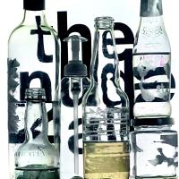 Multiple clear bottles with their labels ripped off sit in front of a screen with text reading, "the nude 4 all".