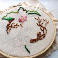 canvas on embroidery hoop with pencil sketch of mayflowers partially filled in with glass seed beeds in pink, green, and brown colours