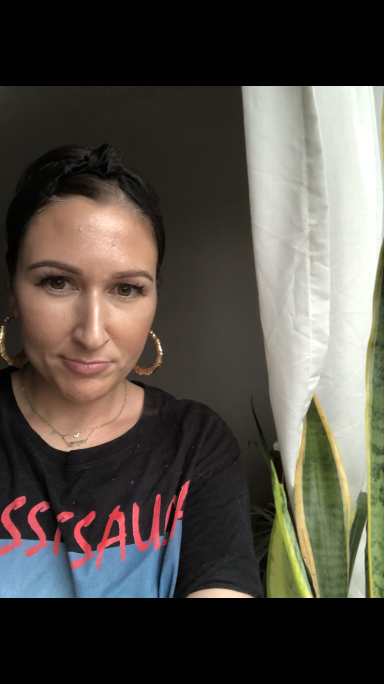 portrait of Ellyn Walker, a light skinned woman looking at the camera with gold hoop earrings in a black tshirt next to a snake plant