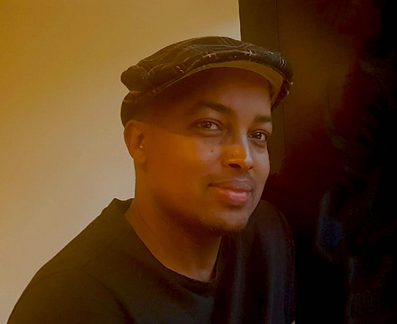 Portrait of Abdi Osman, a dark-skinned man wearing a soft cap and a black t-shirt looking at the camera. 