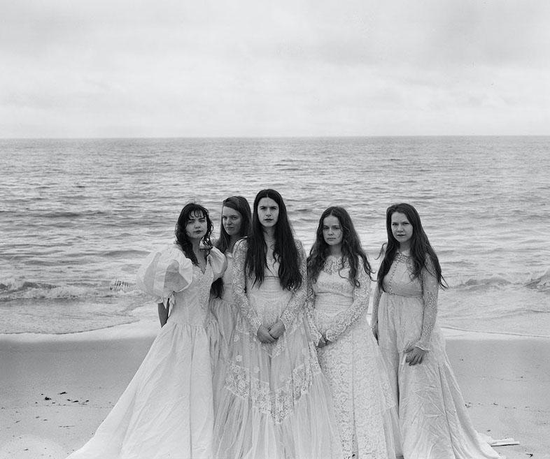 Five women standing in front of the ocean wearing wedding dresses