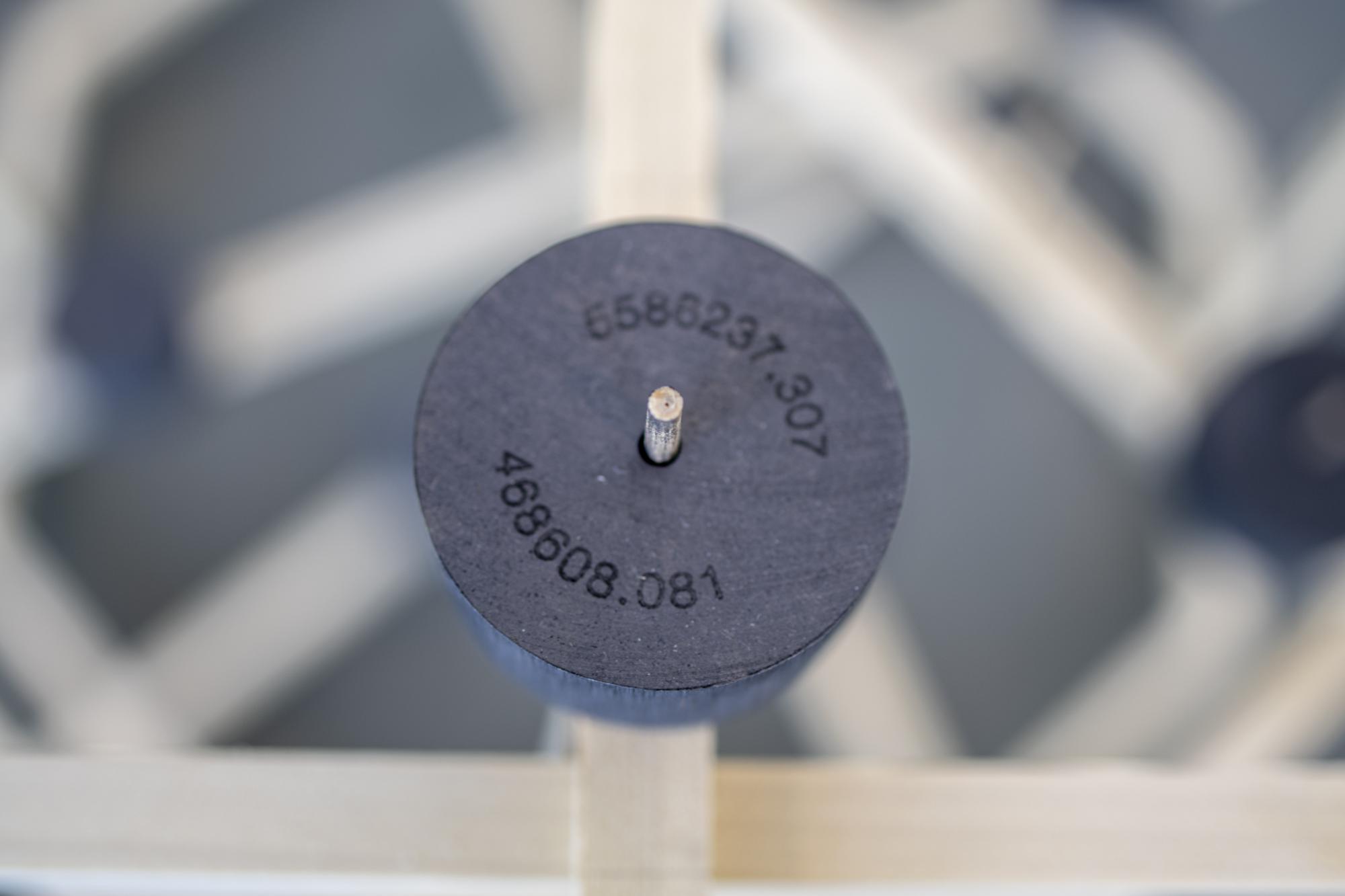 A close-up view of a round graphite block on a wooden structure composed of interlocking sticks. Engraved on the graphite block are latitudinal and longitudinal coordinates that identify the location of a site of difficult history.