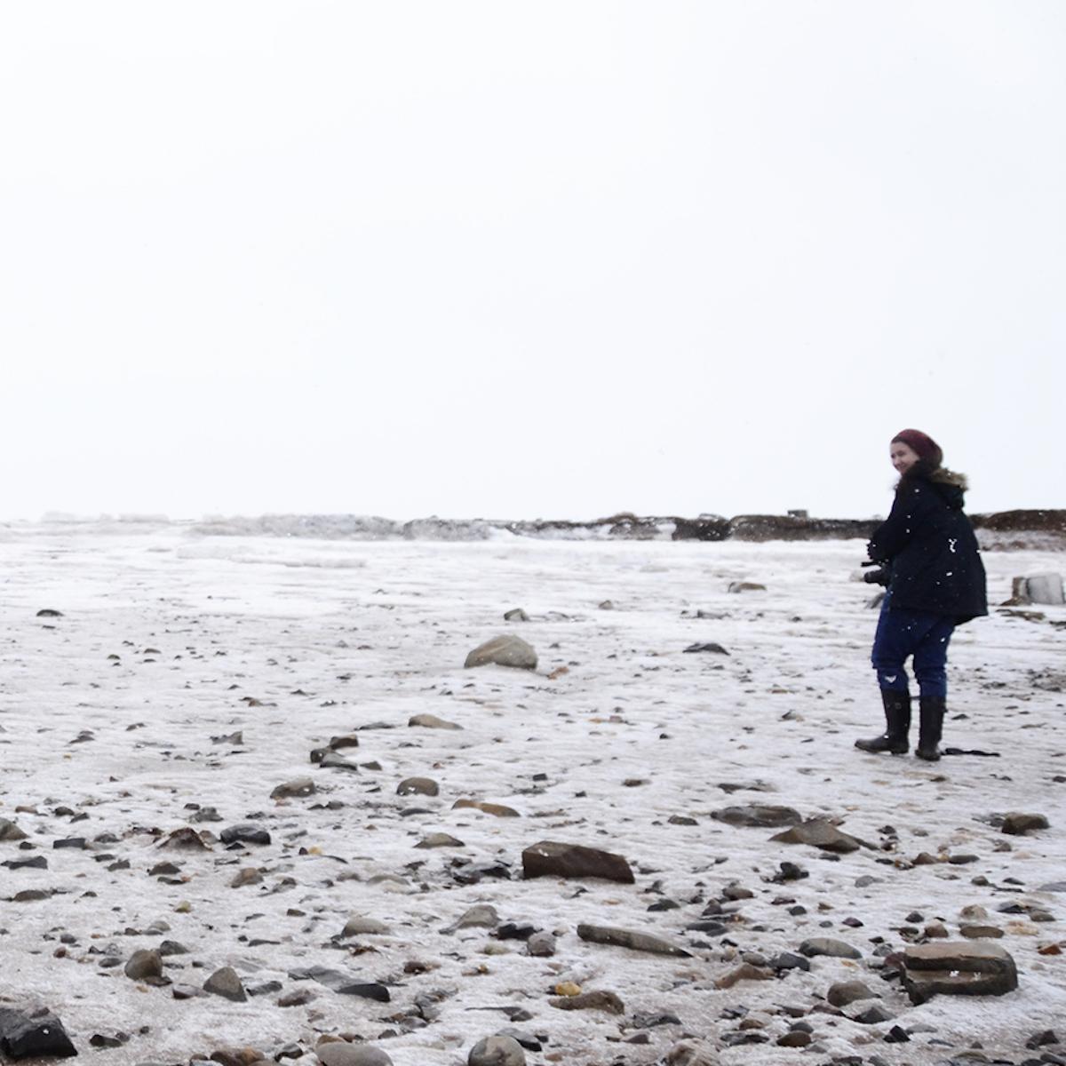  person standing in the snow. They're by the ocean, and the ground is rocky. The person is on the right side of the image, looking back towards the camera. They have olive skin and dark brown hair, and wear a red beanie, black jacket and boots, and blue jeans.