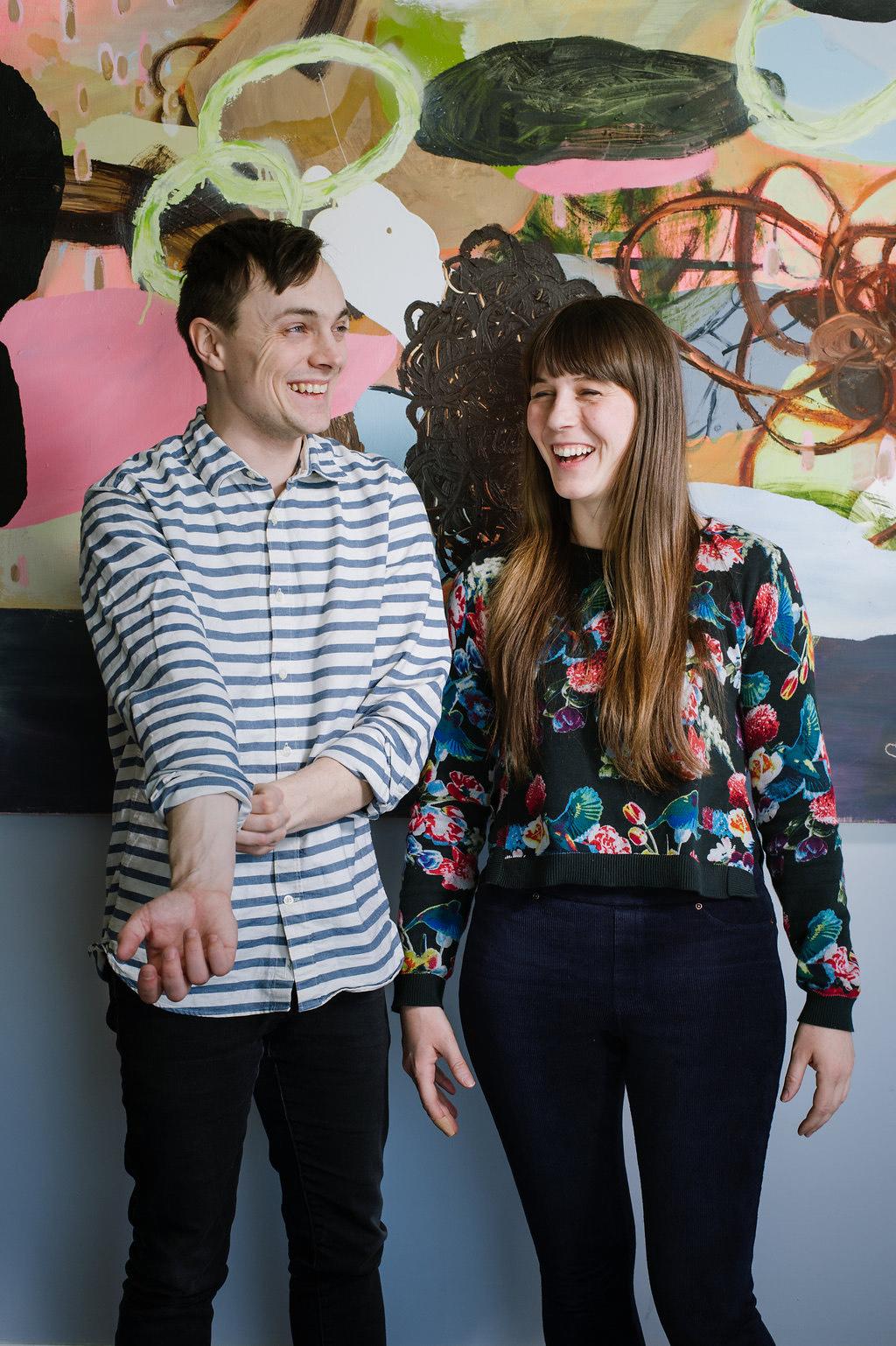 A white male and female laughing looking off camera. An abstract colourful painting is behind them