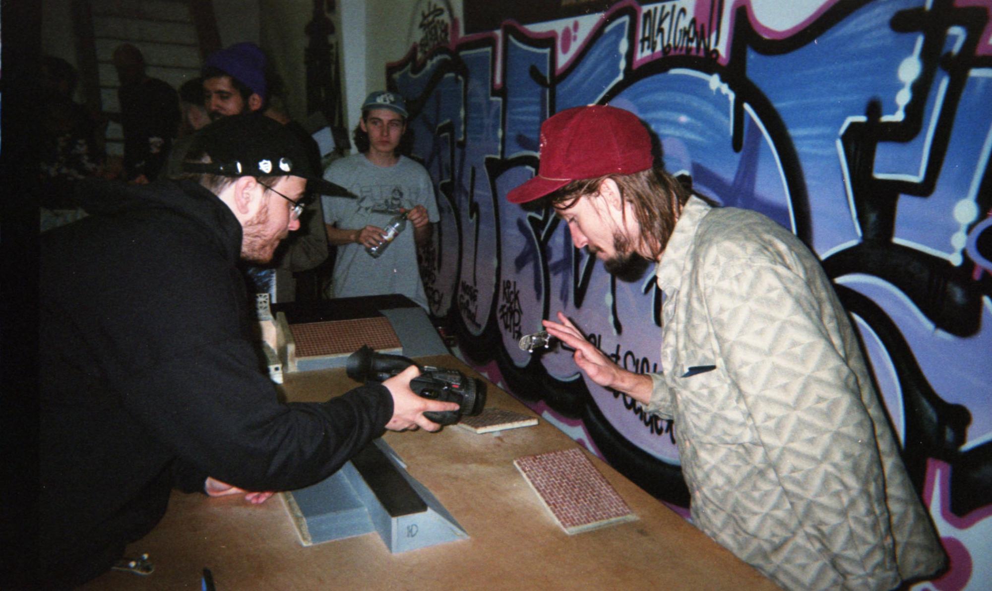 Person wearing red hat and grey coat uses miniature skateboard to do a trick in a miniature skatepark, while another man in black hat and black sweater films.