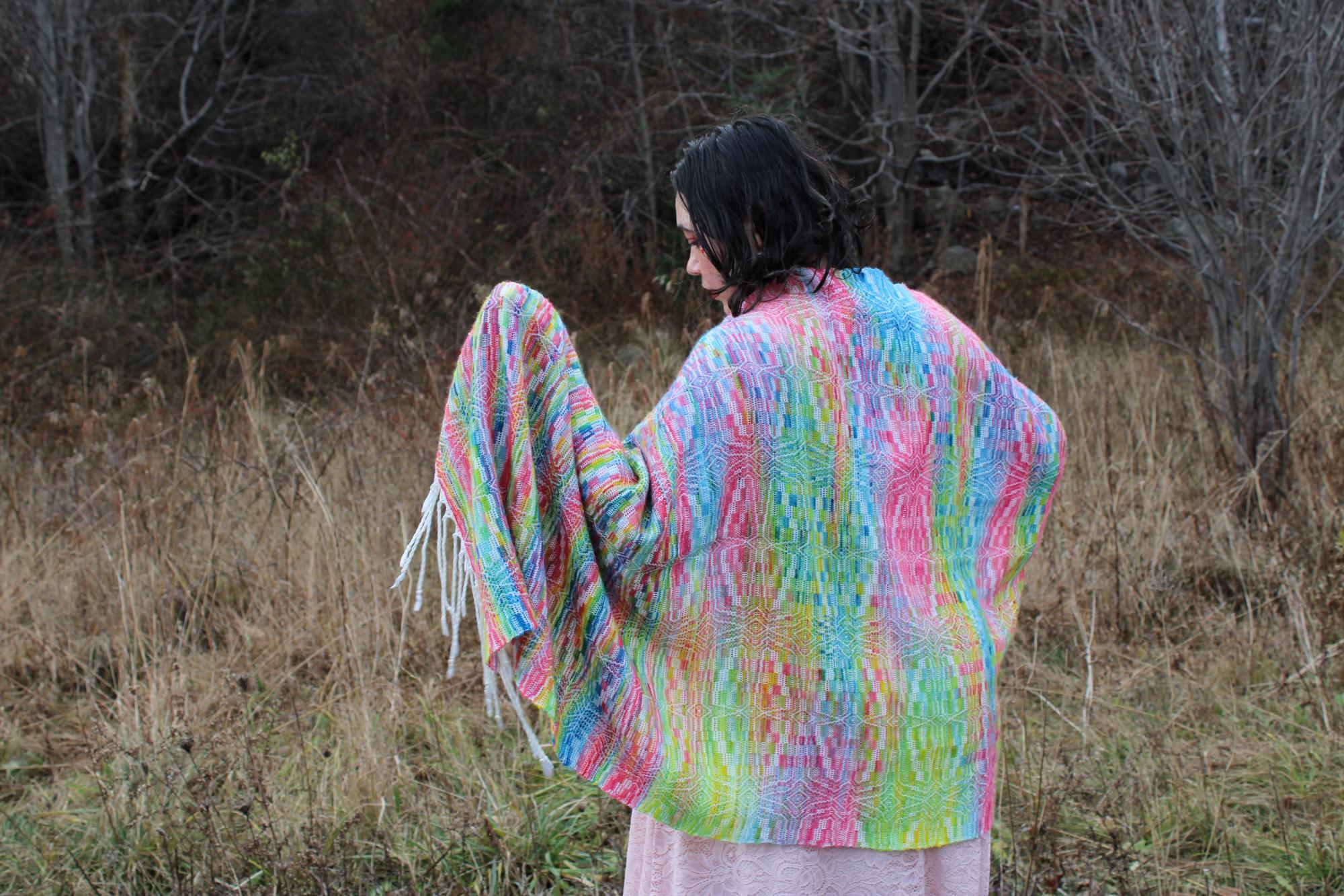 Person stands with rainbow floral shawl facing forest, hand outstretched, looking at part of shawl draped over hand.