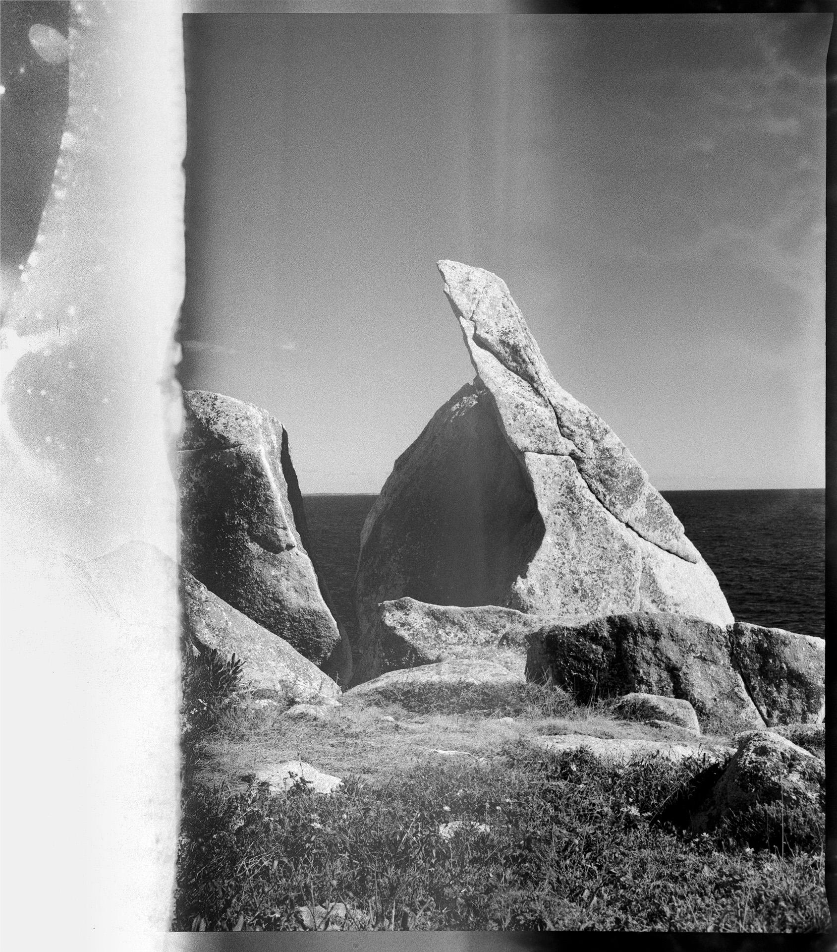 Two large boulders in front of the ocean, that look as if they were split from one boulder. The image is in black and white film and there are fluid looking distortions on the surface of the film. On the left side of the film the image is white with grey drip marks.
