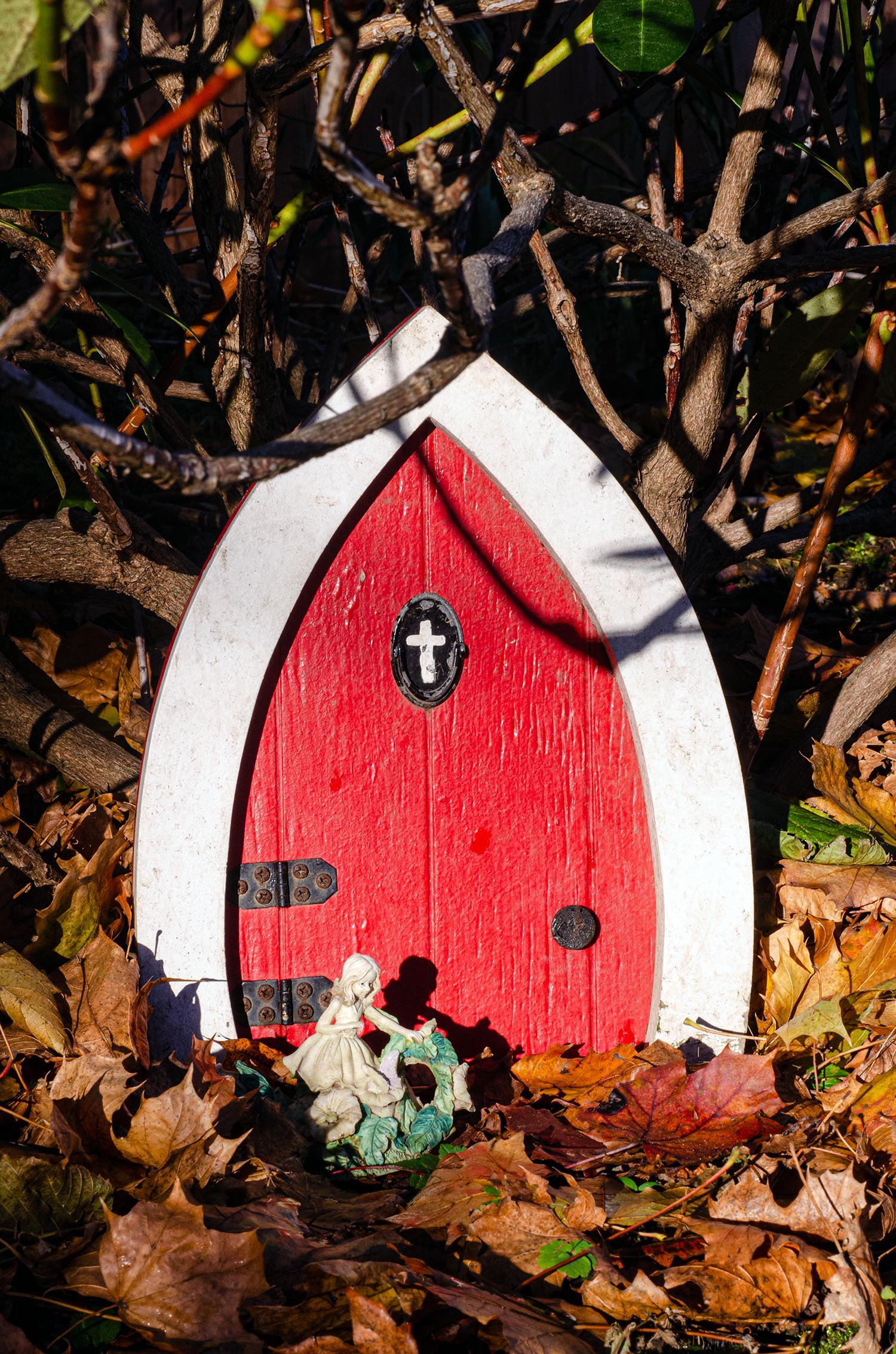 An oval-shaped, wooden, red door with thick white trim sits on a carpet of leaves beneath bare branches of shrubbery. Black metal hinges, a black doorknob, and a flat black oval door knocker with a white painted cross adorn the door. A small figurine is situated in front.
