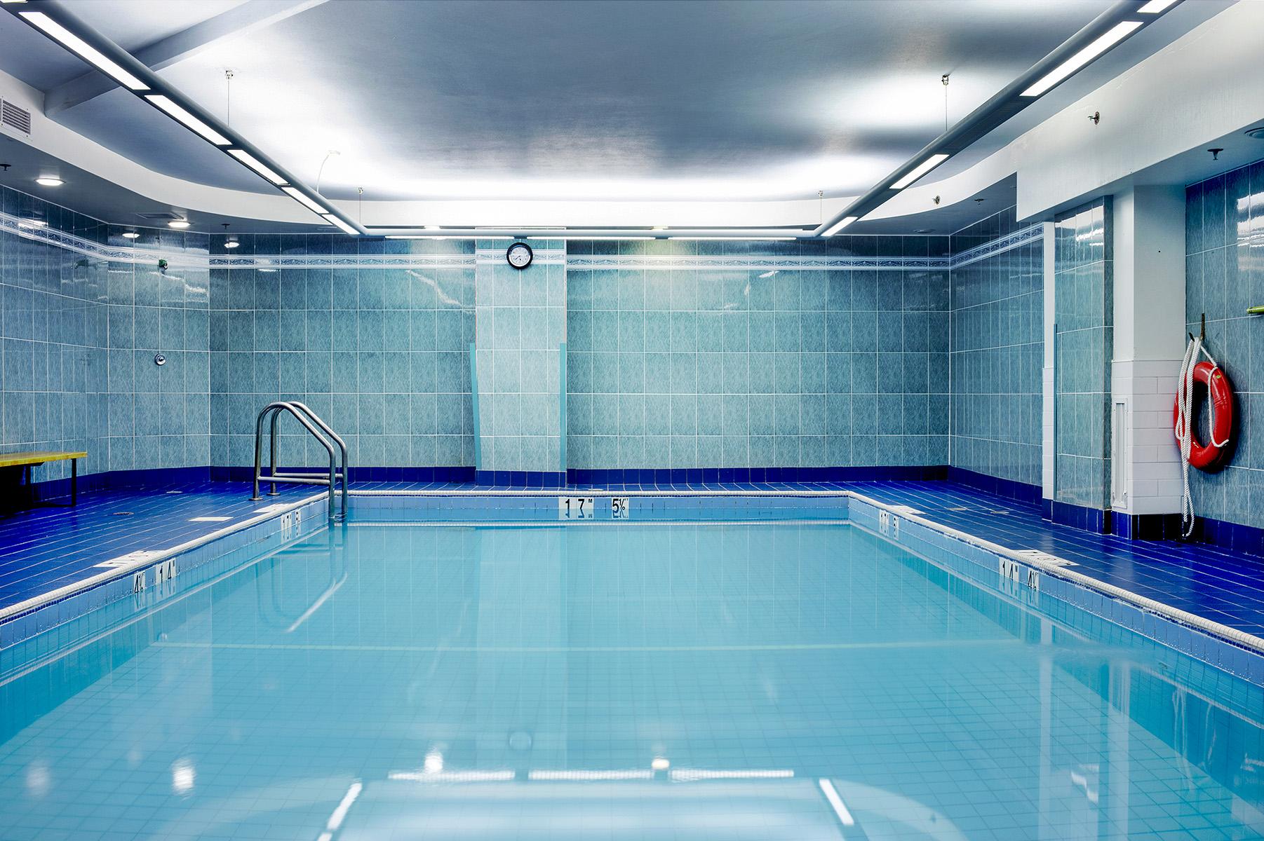 An indoor hotel swimming pool, with blue and turquoise tiles on floor and walls. Two long headlamps hanging from the ceiling, a black clock on the center wall, a wood bench on the left side, and an orange lifebuoy hanging on the right wall.