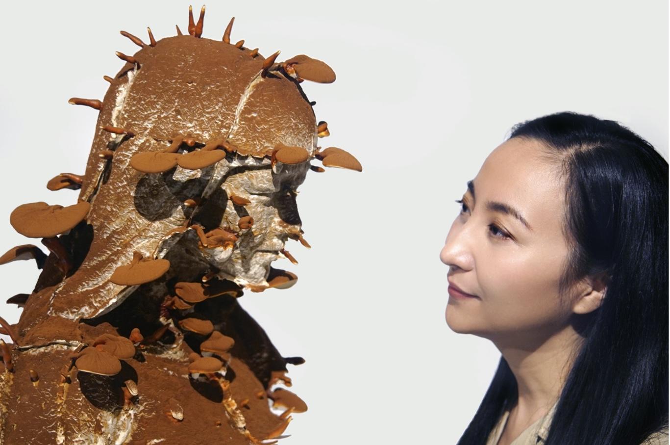 On the left, sculptural work of a figure with various sized circular pieces attached to its head, neck and shoulders. The sculpture is covered in medium-dark reddish brown rust. On the right, a person stands in front of the sculpture, looking into its eyes. 
