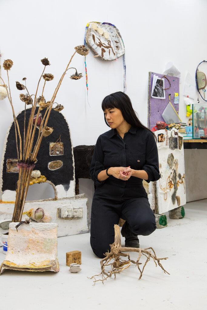 Medium-dark skinned person crochet in centre of frame, in a predominantly white studio space. The person is looking to their right at a mixed media sculpture.