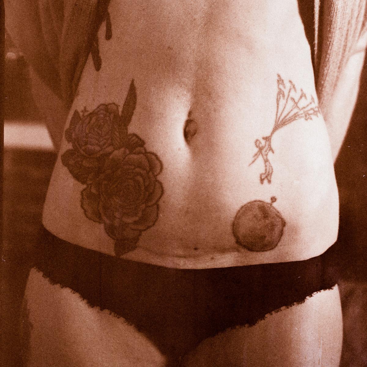 Sepia tinted closely framed photo of an abdomen with tattoos and a cesarian section scar. 