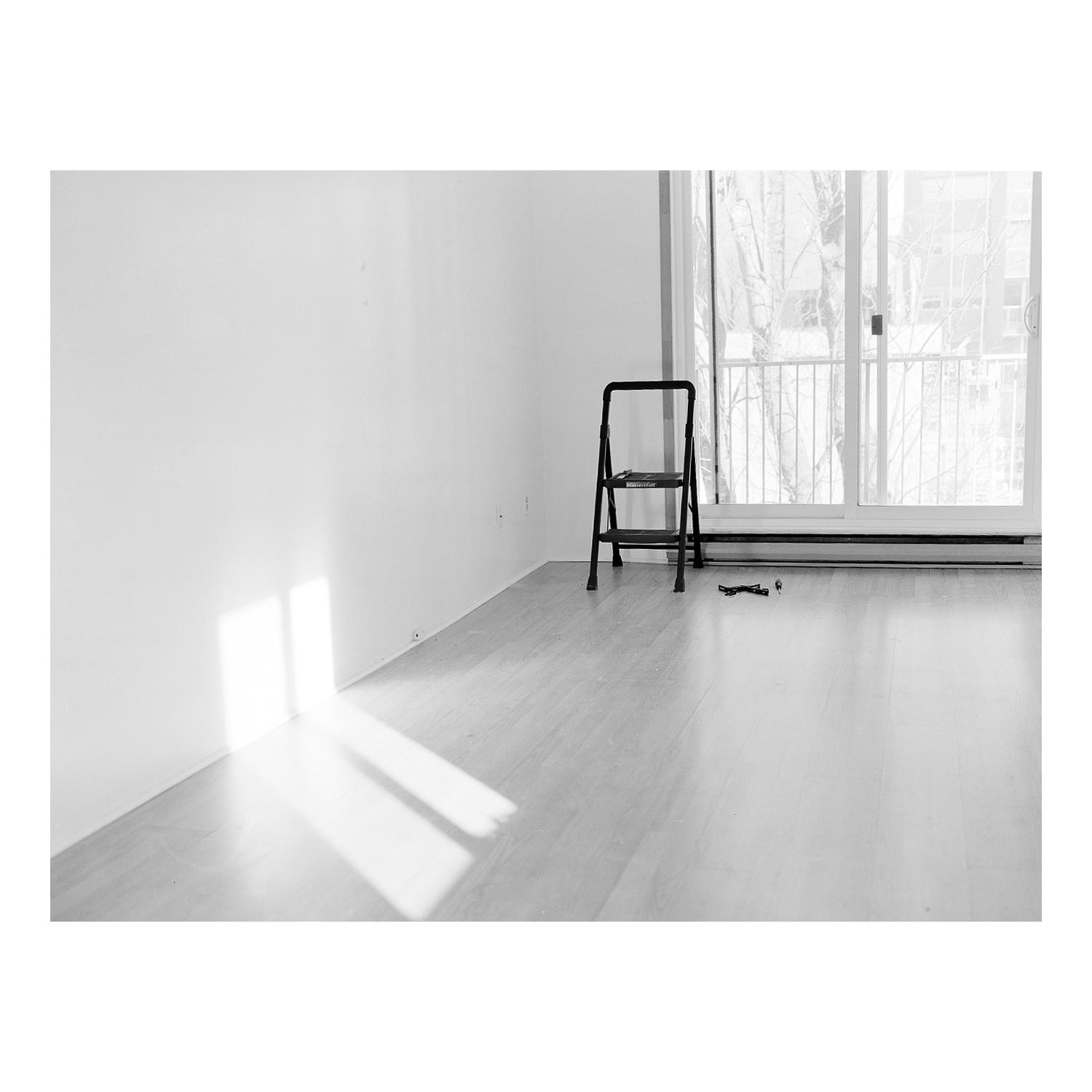 Black-and-white image of an empty room. A stepladder sits near a sliding glass door. Light reflects off the wall and floor.