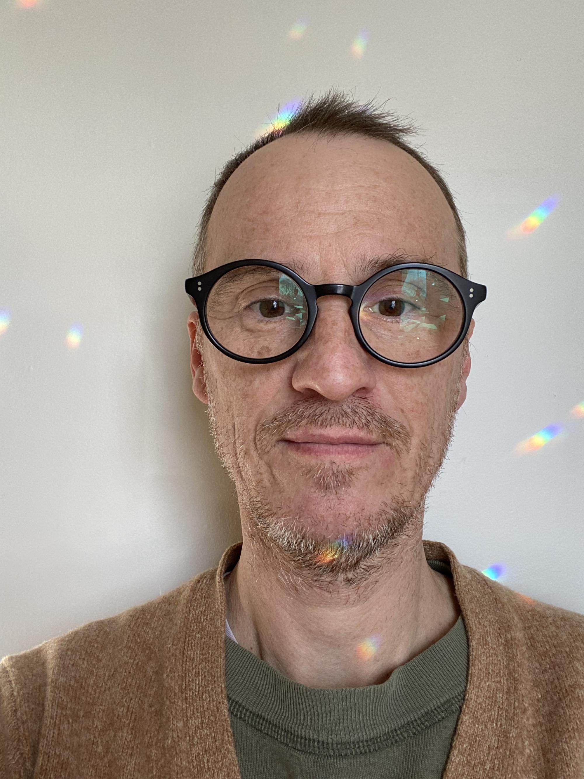 Light skinned person wearing thick framed glasses, green shirt and brown cardigan. Beams of light cast upon person and background.