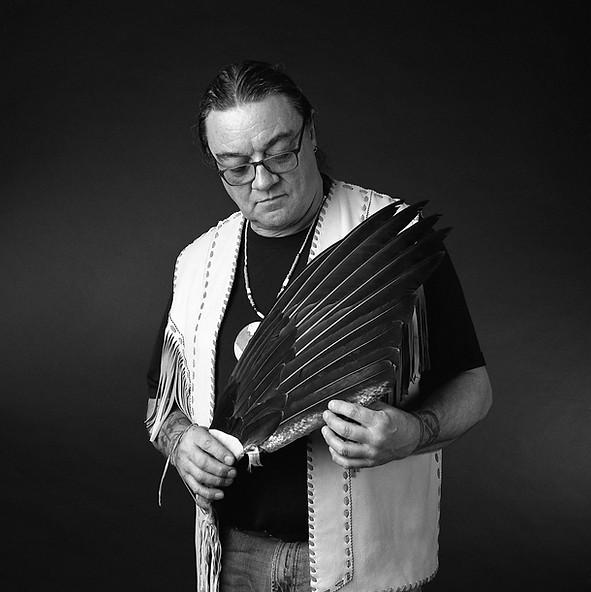 Black and white photography of artist standing in centre, wearing glasses and holding feather piece.