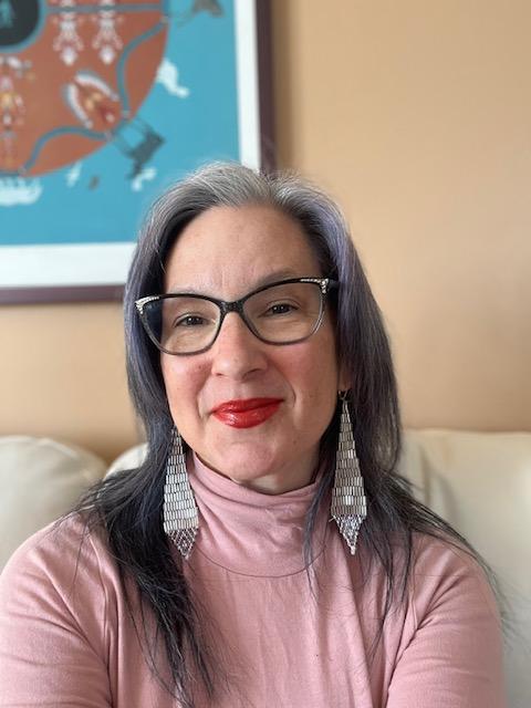 Photograph of Cathy Mattes smiling at the camera. She is wearing a light pink turtleneck, silver beaded earrings, red lipstick, and black glasses. Her light brown and gray hair is down and falls past her shoulders.