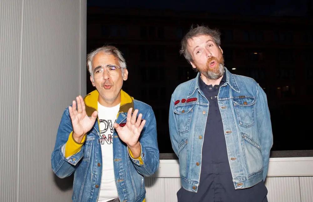 Peter Morin and Jimmie Kilpatrick stand next to one another with their mouths open in song. Both are wearing denim jackets. Peter has his hands outstretched and Jimmie has his hands clasped behind his back.