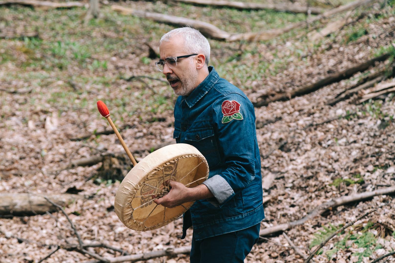 Peter Morin stands in the forest wearing a denim jacket with a beaded rose patch on his shoulder. He holds a rawhide hand drum in one hand and a drumstick in the other, his mouth open in song.