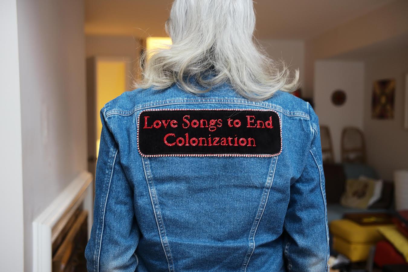 Photograph of a person with shoulder length gray hair standing with their back facing the camera. They are wearing a denim jacket with a large patch spanning the width of their shoulder that reads "Love Songs to End Colonization" beaded in small red glass beads.