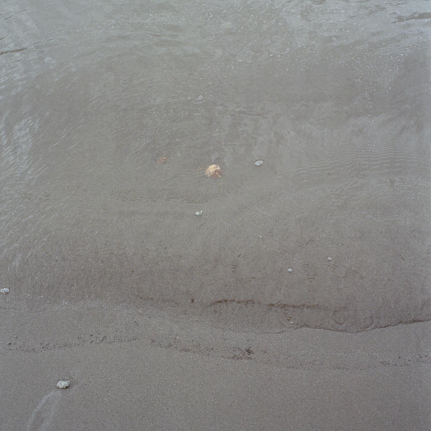 Close up photograph of grey sand with pebbles amongst the sand and water lightly washing over it.