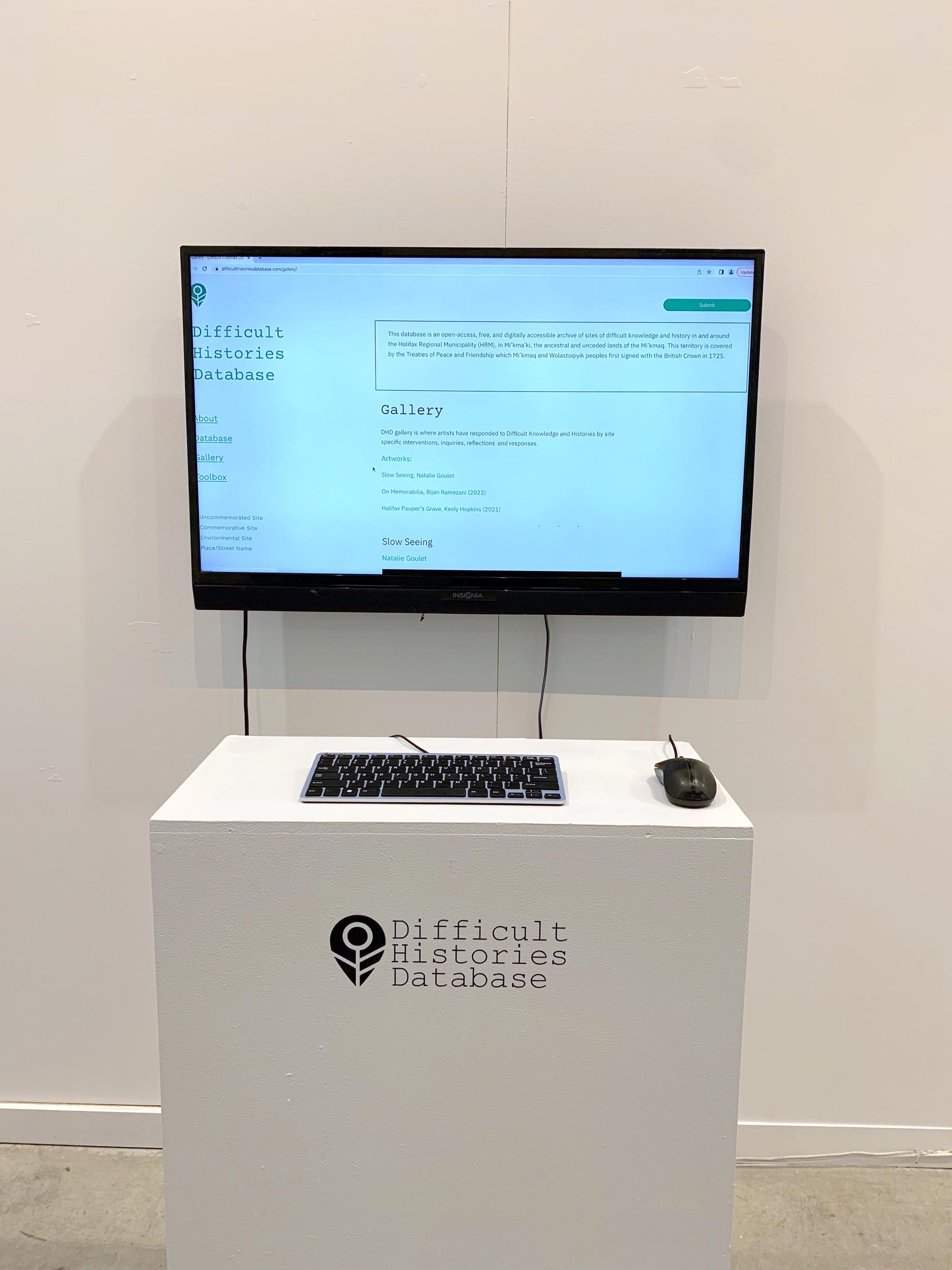A large screen displays the Database website, there is a keyboard and mouse on a plinth in front of the screen with the DHD logo printed in black vinyl on it