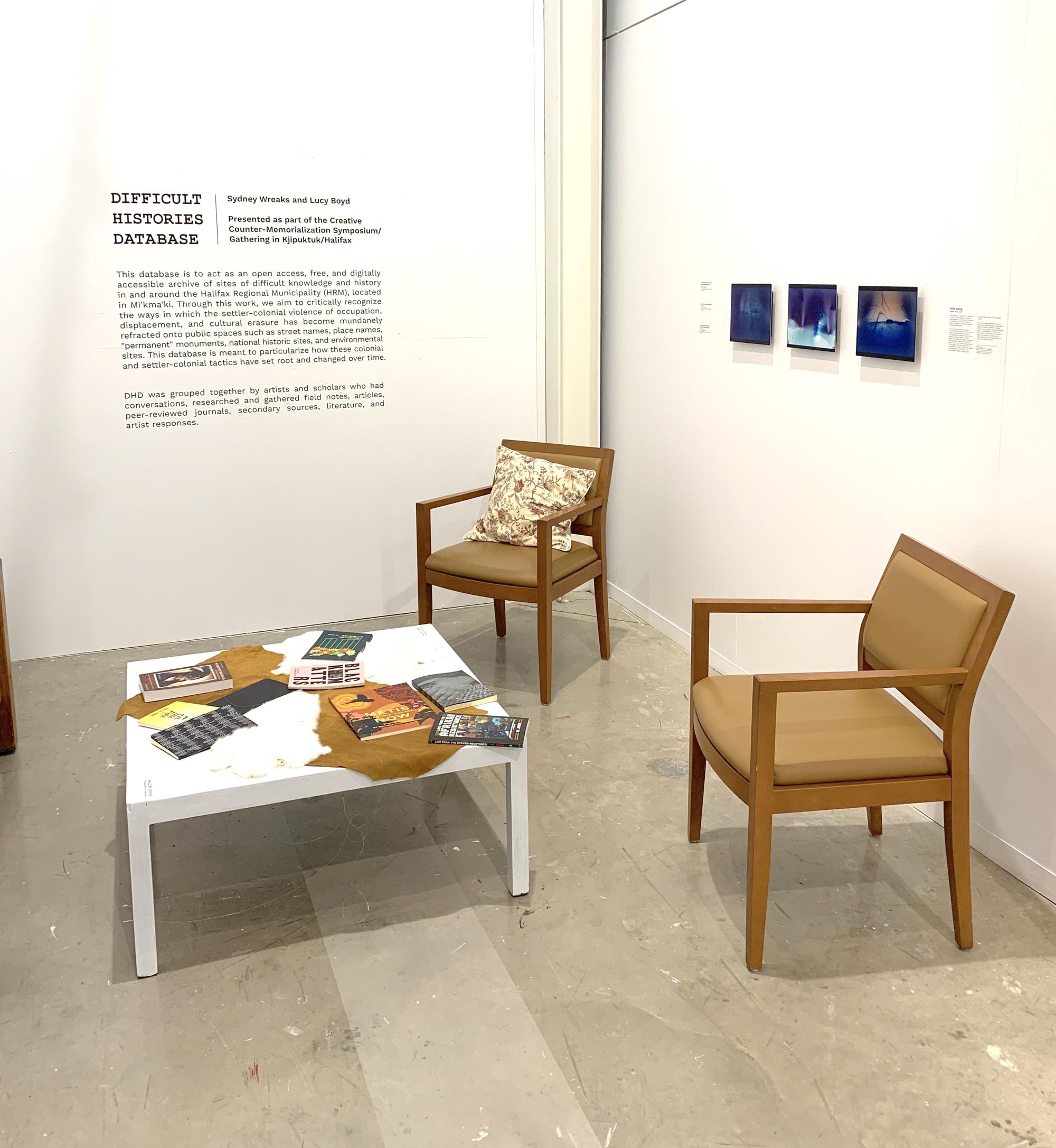 two chairs surround a white coffee table with various books, 3 artwork prints are on the wall behind the chairs, exhibition black vinyl text on adjoining wall