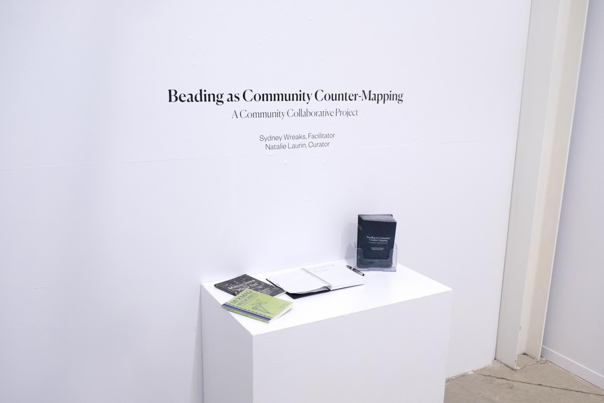 plinth with guest book and exhibition catalogue underneath wall with black vinyl text showing exhibition title