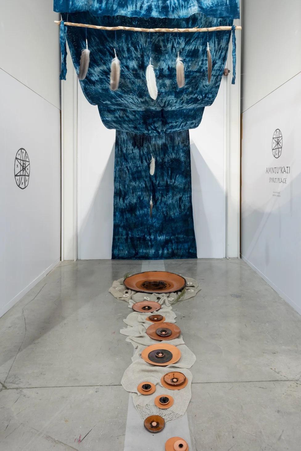 photograph of installation with ceramic bowls of varying sizes in the centre of the floor leading to a large blue tapestry on the back wall that continues up across the ceiling