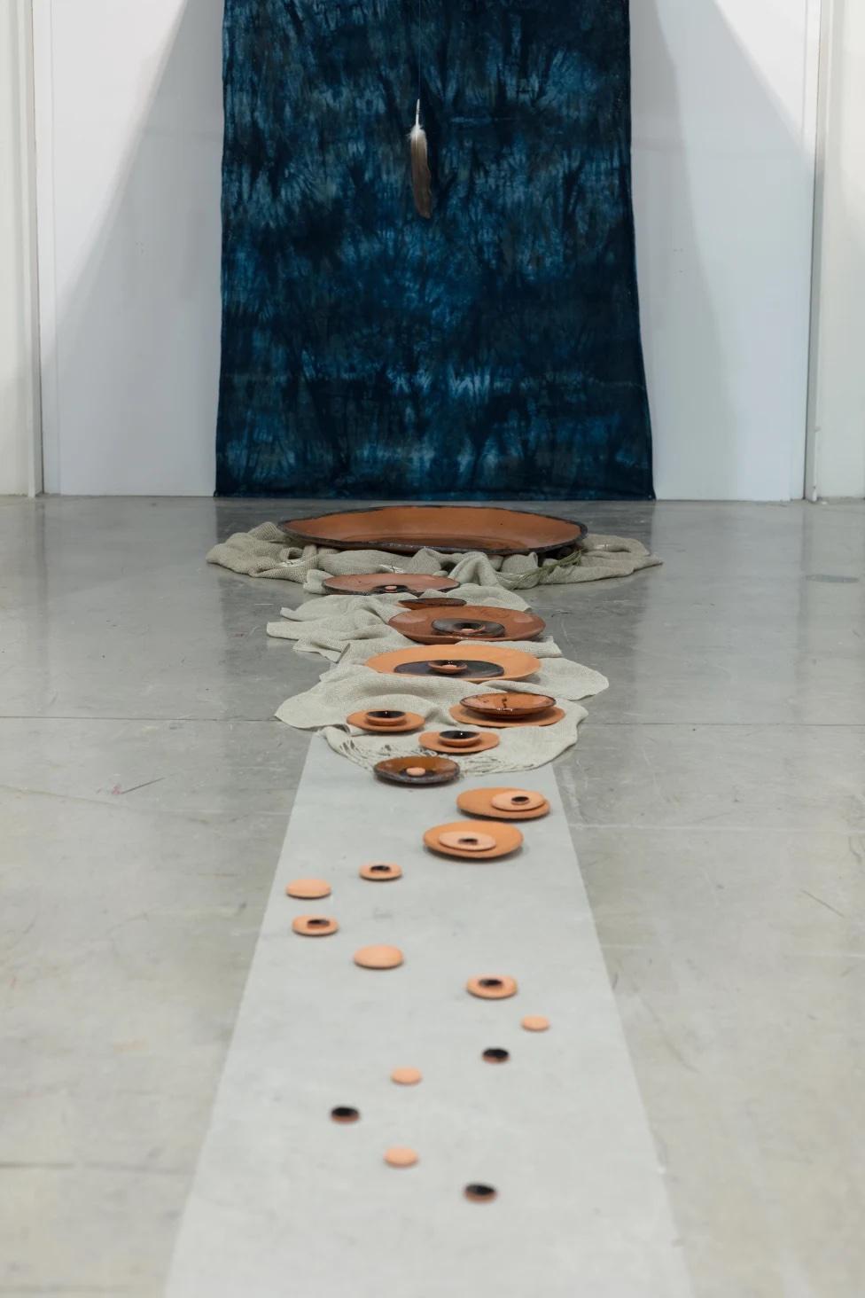 photograph of installation with ceramic bowls of varying sizes in the centre of the floor leading to a large blue tapestry on the back wall that continues up across the ceiling