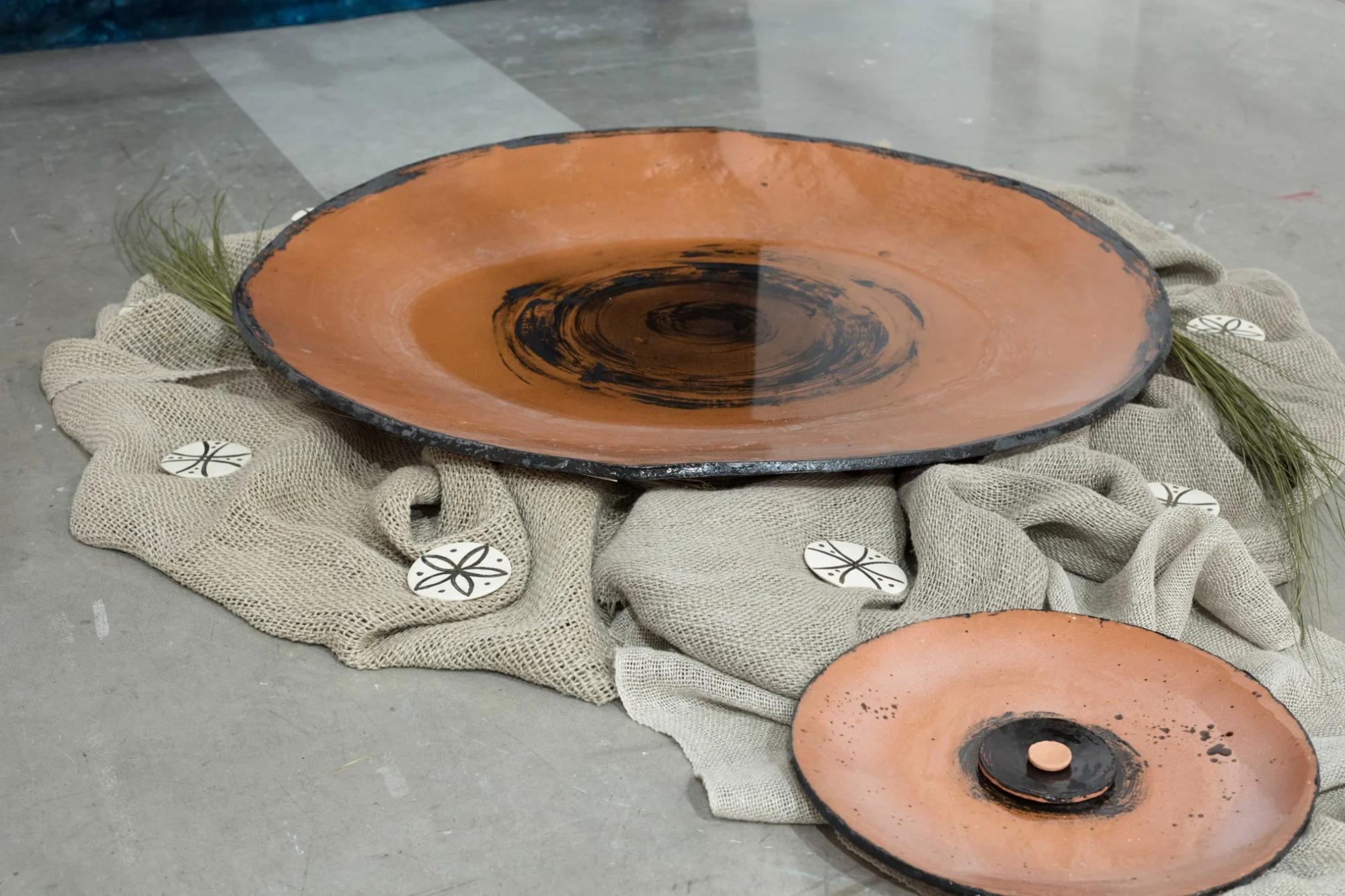 close up of two ceramic dishes on the floor placed onto a beige textile. The larger dish has some water in it and is surrounded by smaller white, round ceramic pieces with dark coloured designs on them 