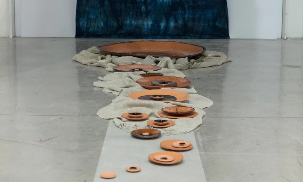 ceramic dishes varying in size sit on a gray concrete floor leading towards a wall with a large blue textile hanging on it