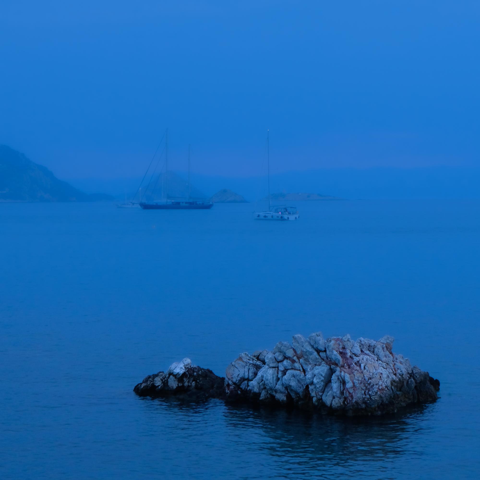 Photograph of a hazy blue sea with rocks in the foreground and large sailboats in the background