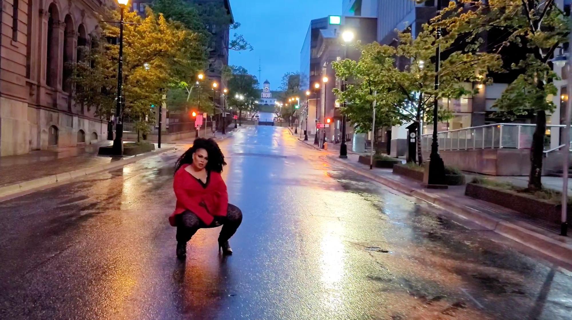 A person wearing black leggings and high heels with a bright red blazer and long black curly hair crouches in the middle of the street, looking at the camera. The sky is a dark blue and the street glistens with rain. The Halifax Citadel Hill clock tower can be seen in the background at the top of the street. 