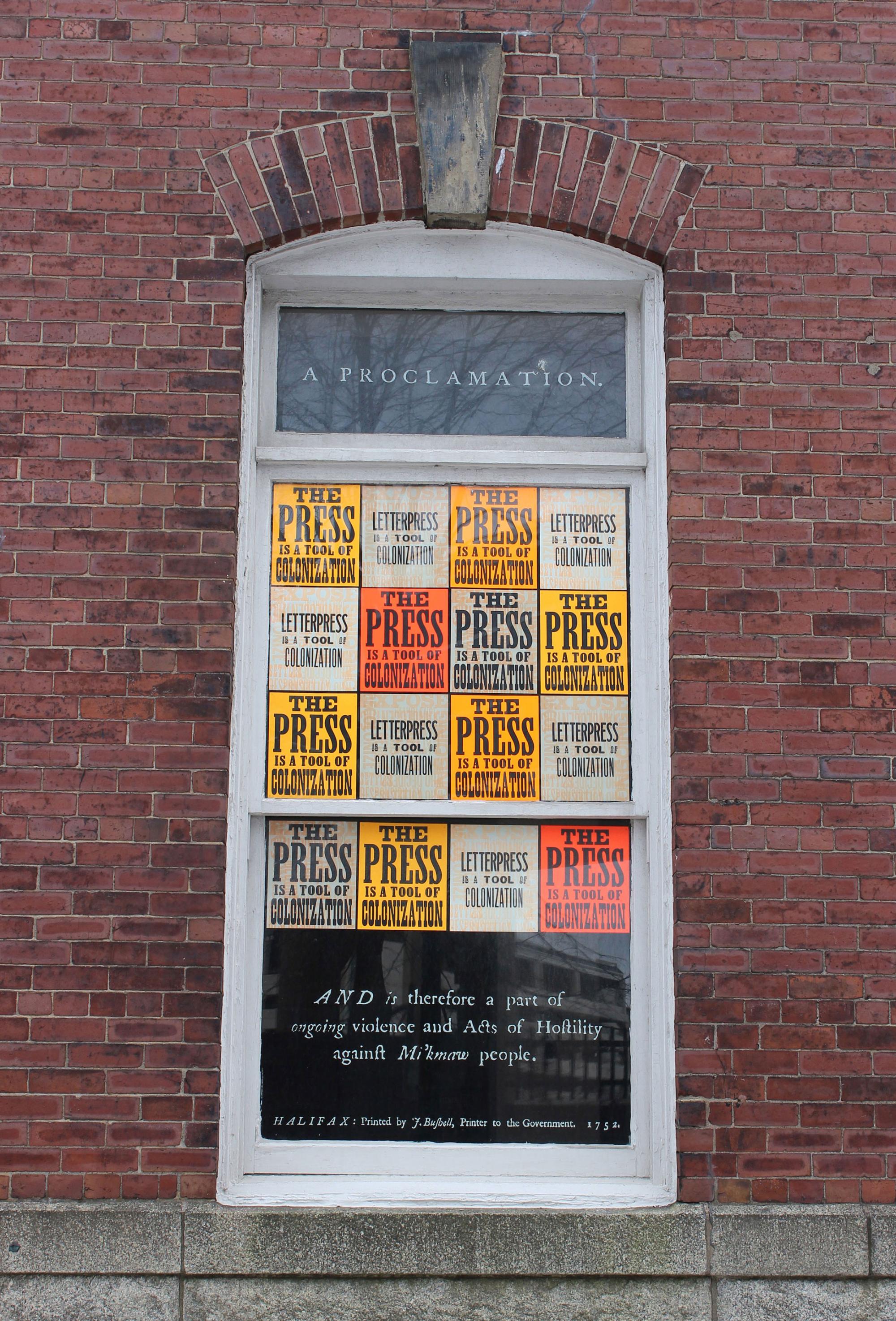 Neon orange letterpress prints hang in the window of a brick building. Prints read "Letterpress is a tool of colonization.”