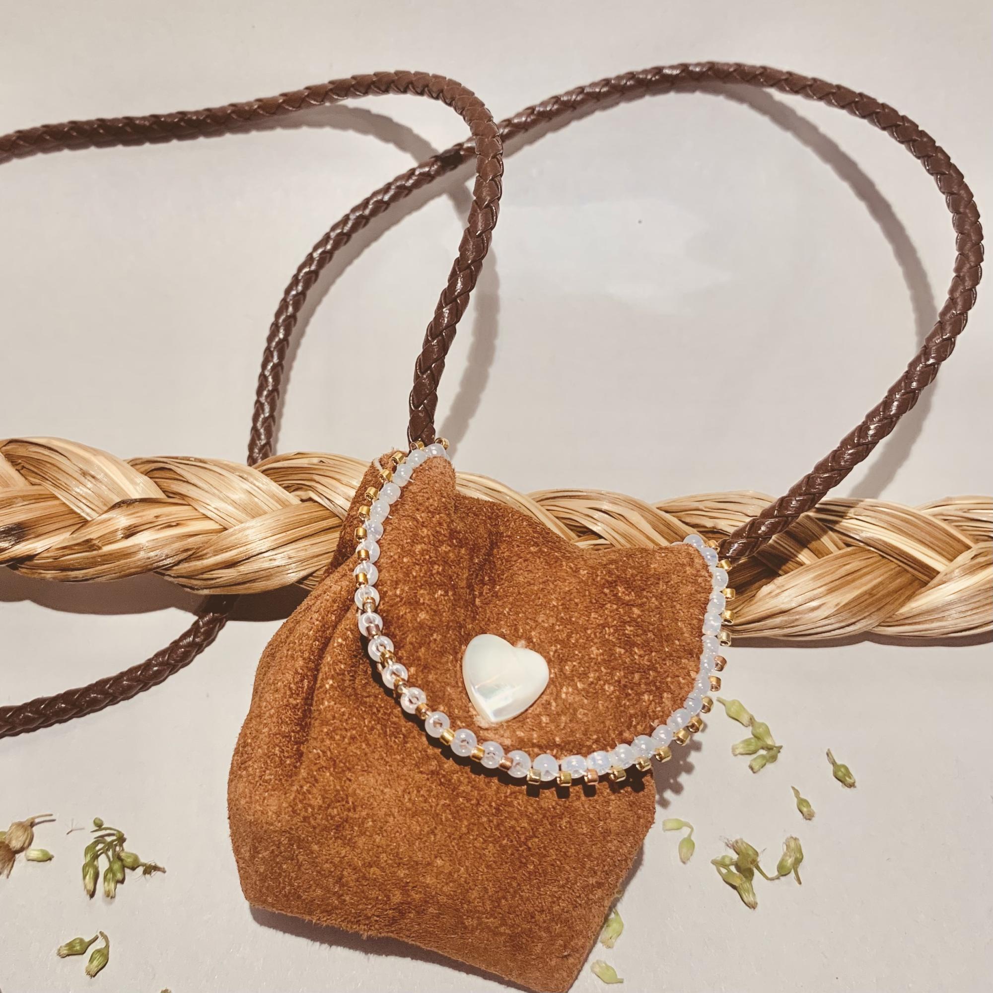 [ID: "Brown leather medicine bag beaded with white beads laying on top of braided sweetgrass and surrounded by green seeds."]