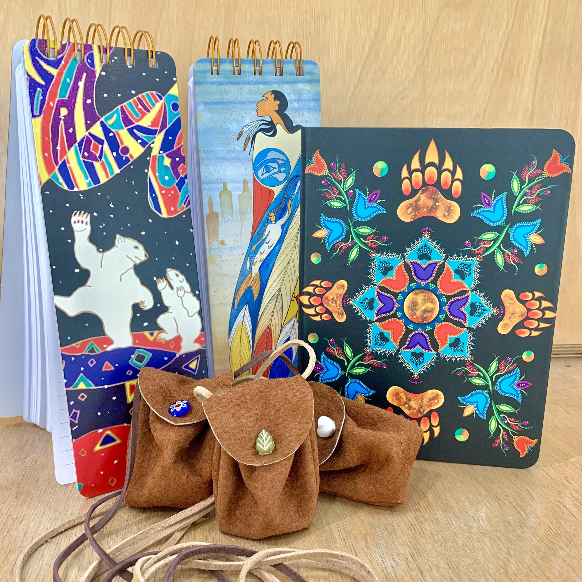 three leather medicine bags sit atop a wooden surface in front of 3 notebooks with colourful designs on them created by Indigenous artists