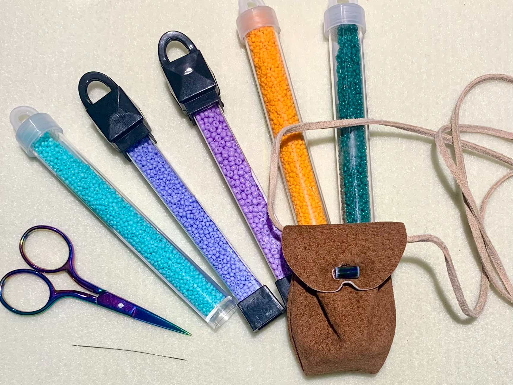 [ID: "Blue, purple, orange, and turquoise beads each in a cylindrical tube, a small pair of purple scissors, and a sewing needle surrounding a brown leather medicine bag. The background is a flat white surface."]