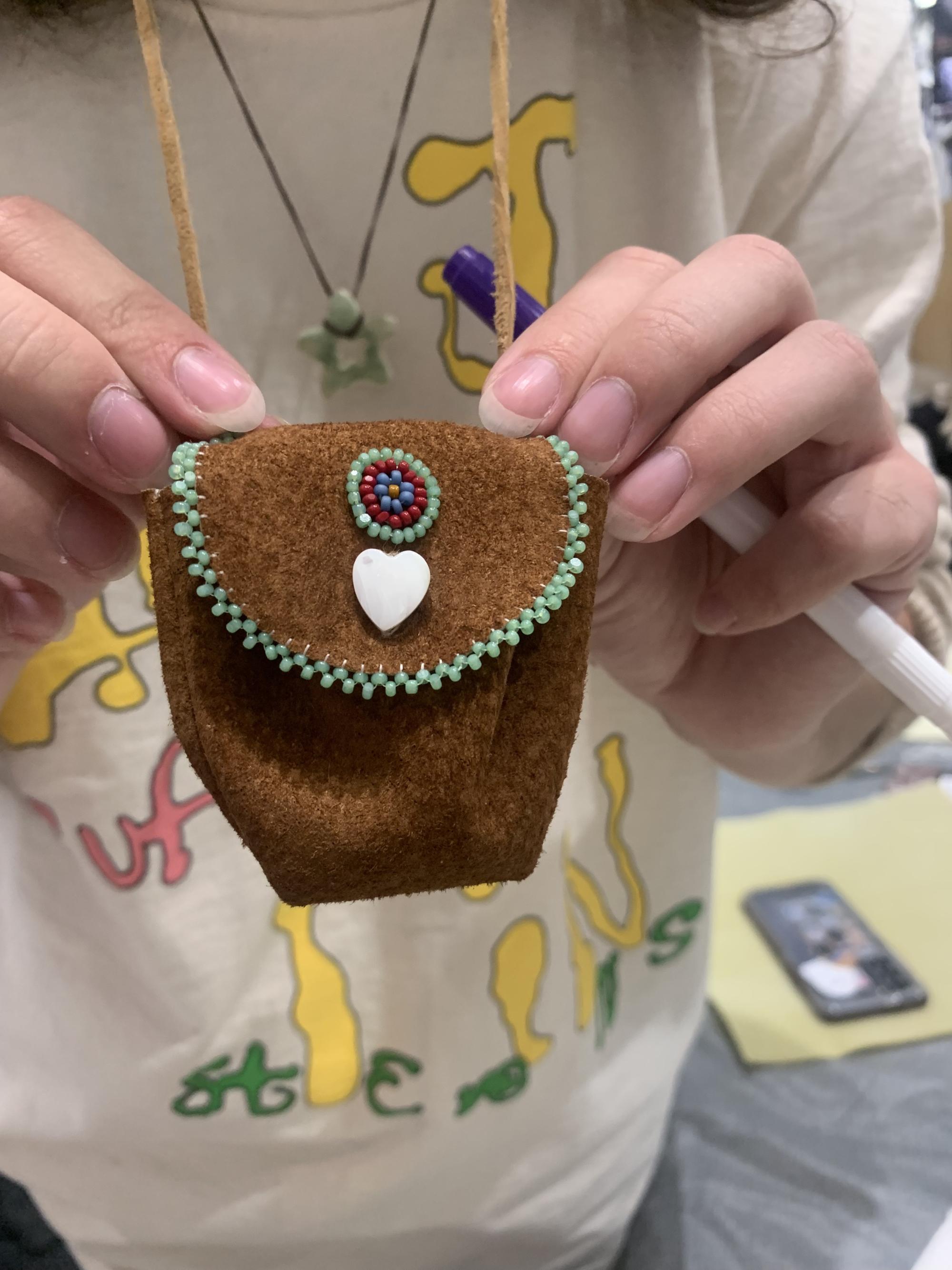 [ID: "Workshop participant holding up the beaded medicine bag that is hanging from their neck. The light brown leather medicine bag is beaded along the edges with green and white beads, has a white heart button closure, and a circular beaded detail."]
