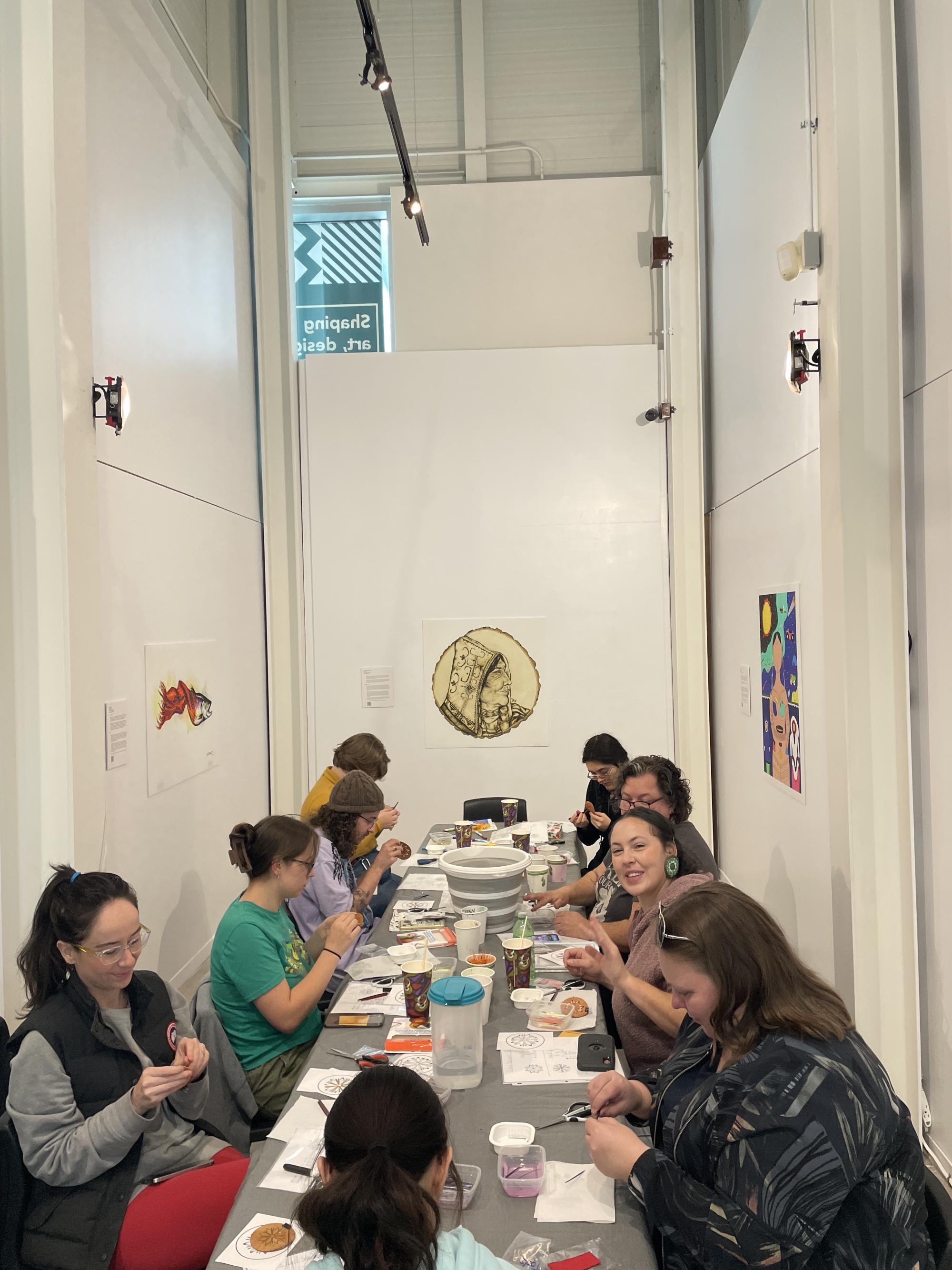 [ID: "Porcupine quill workshop participants working on their medallions at a long table in the Treaty Space Gallery."]