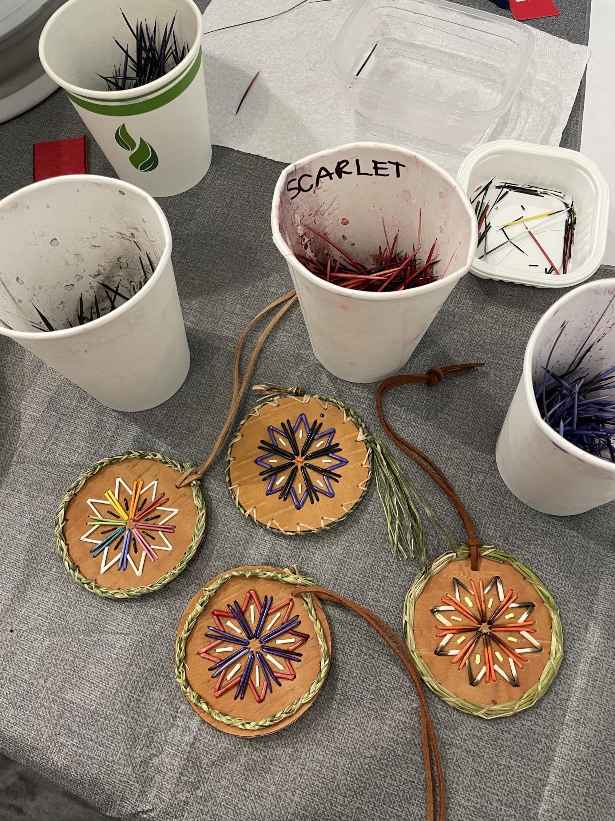 [ID: "Four colourful porcupine quill medallions made by workshop participants with birch bark, sweetgrass, and dyed quills."]