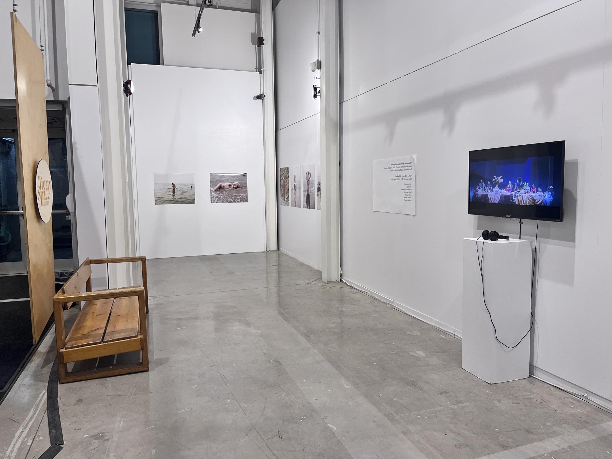 [ID: "Wide shot of the NSCAD Treaty Space Gallery. In the foreground there is a wooden bench, a TV mounted to the wall, and headphones on a plinth. In the background there are various photos by artist Princex Naveed."]