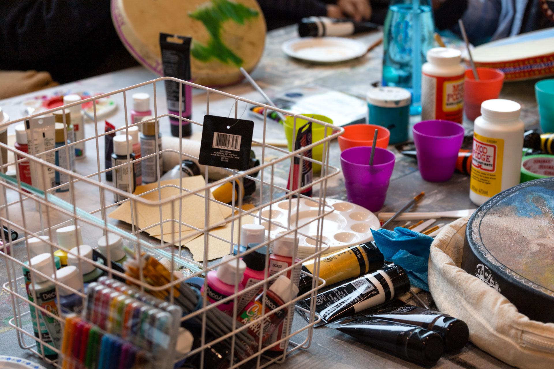 photograph of a table filled with craft supplies like tubes of brightly colored paint, brushes, plastic water cups