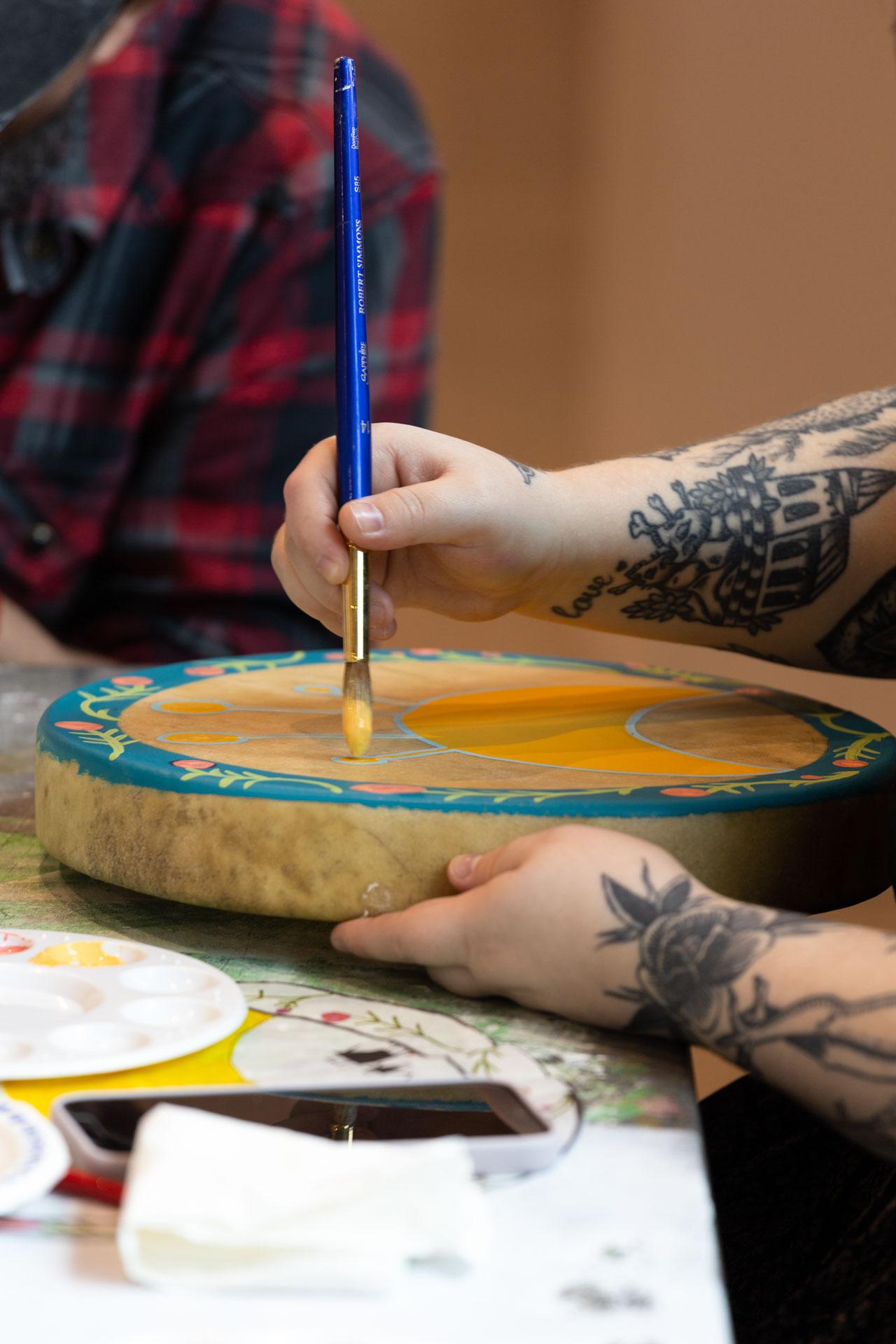 photograph of an artist holding a rawhide drum resting on a table, while brushing a paintbrush with yellow paint onto its surface