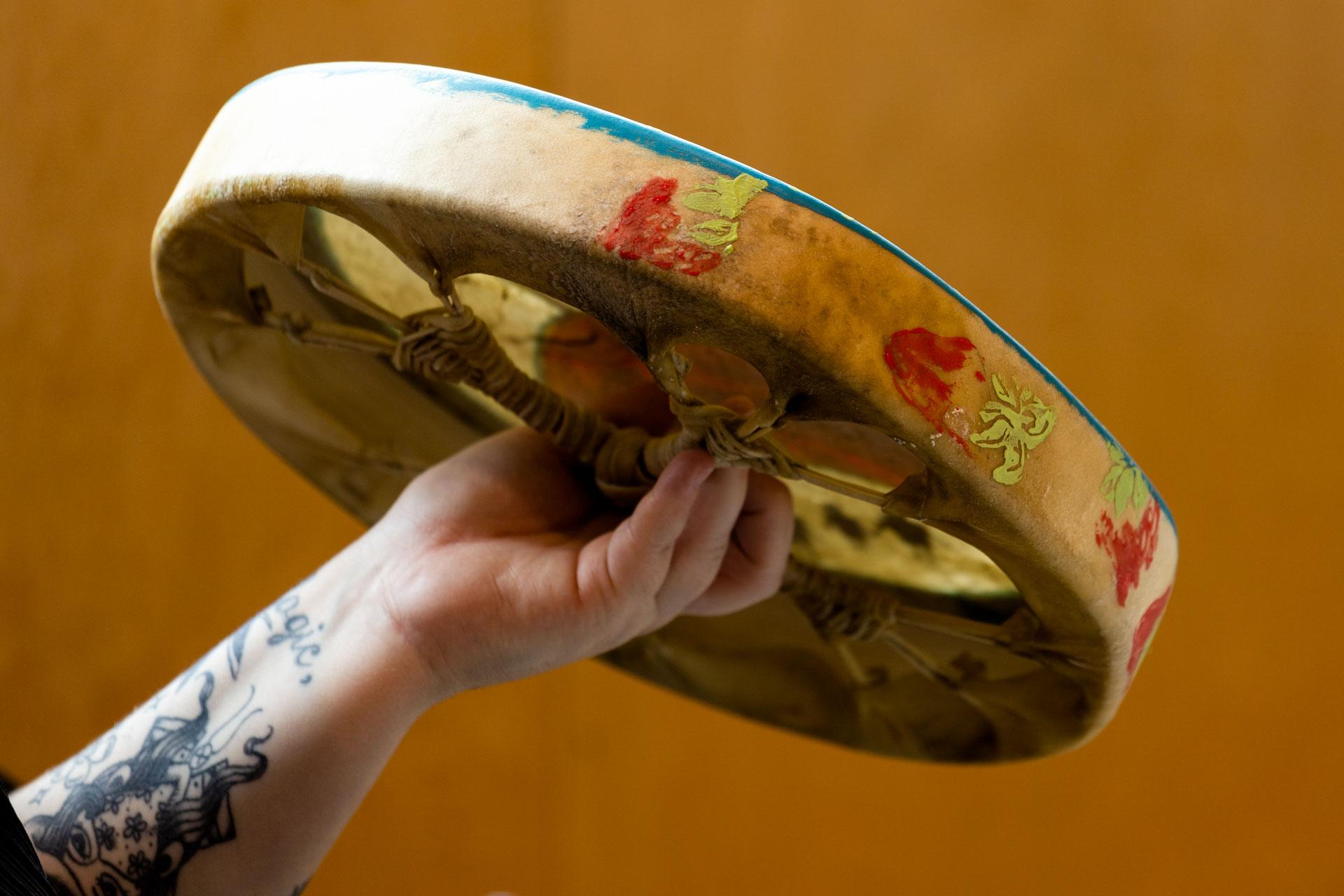 A tattooed arm holds out a rawhide drum, stamped with a strawberry design on its outer edge. The rawhide lacing on the back of the drum is visible.