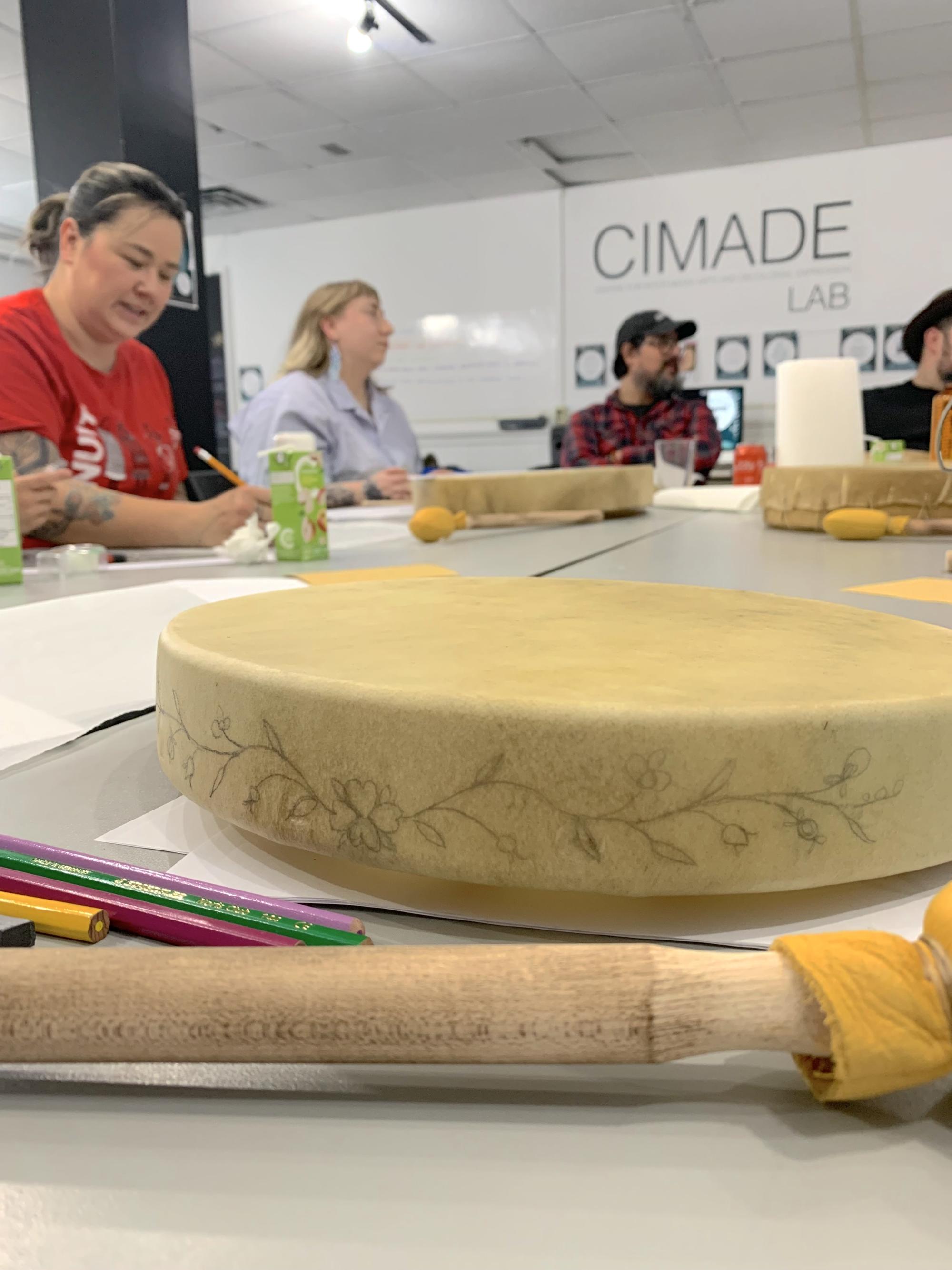 Photograph of a rawhide drum in the foreground, with pencil sketch on its outer edge of a floral design. Workshop participants can be seen smiling in the background