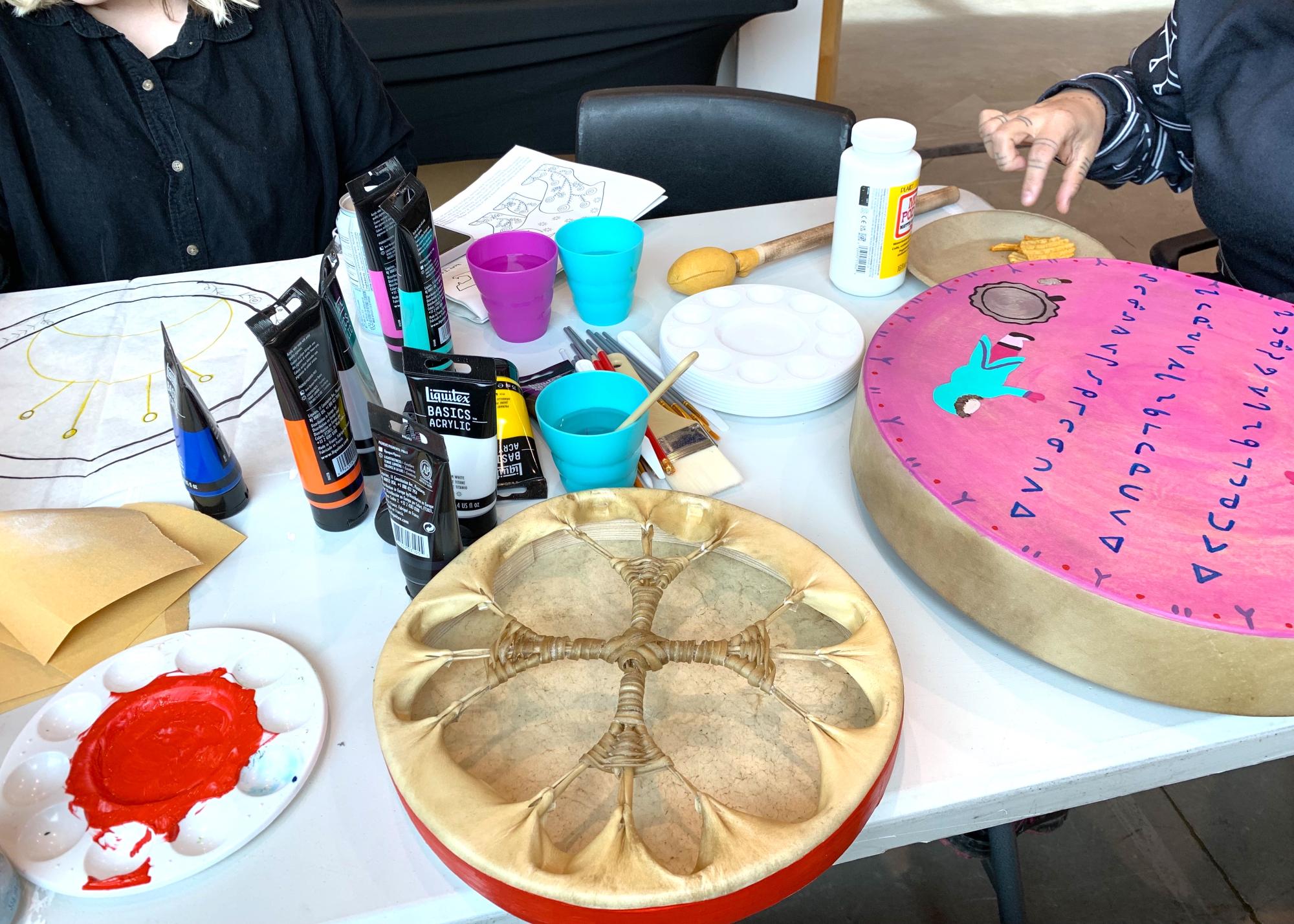 photo of two rawhide drums on a craft table next to tubes of paint and palettes. One is on its face, with the criss-cross rawhide lacing on the back exposed and red stripe around its outer edge. One is painted pink with blue syllabics 